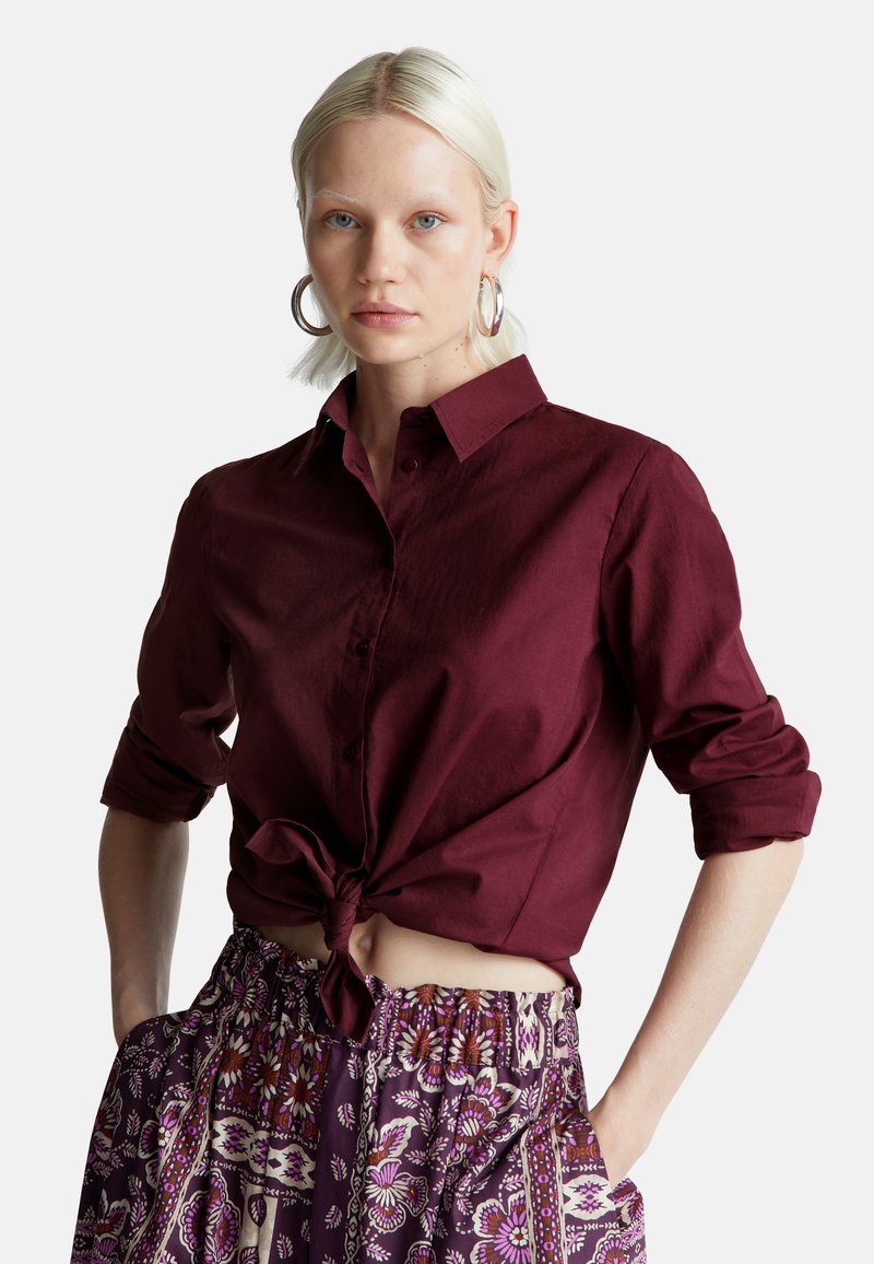 Bordeauxfarbene Hemdbluse mit hochgerollten Ärmeln, die an der Taille gebunden ist, kombiniert mit einem gemusterten Rock mit elastischem Bund, der florale Designs aufweist.