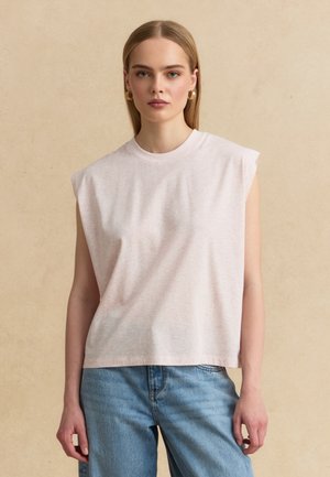 Giovane donna con capelli biondi lisci indossa una canotta beige chiaro con spalle imbottite e jeans di denim blu, in piedi contro un muro beige.