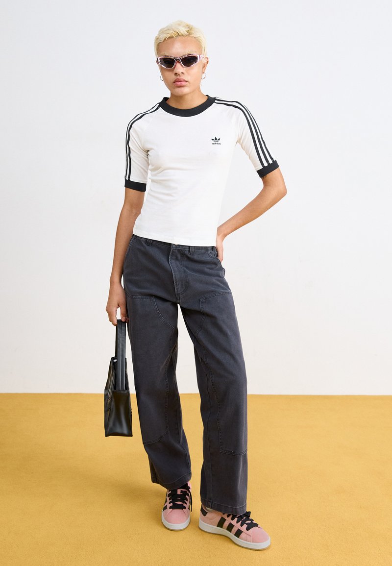 Weißes Kurzarmhemd mit schwarzem Saum und drei schwarzen Streifen an den Ärmeln. Dunkelgraue weit geschnittene Hose und rosa Sneakers mit schwarzen Streifen. Schwarze Tasche.