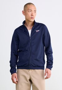Námornícky zipsový kabát s vysokým golierom, dlhými rukávmi, ribbovanými manžetami a logom Reebok na ľavej hrudi. Hladká tkanina bez vzorov.