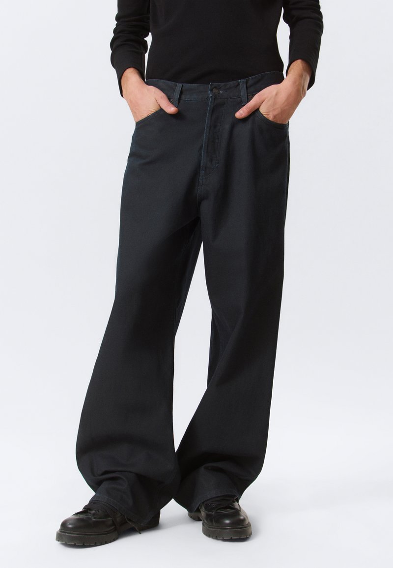 Personne portant un pantalon noir ample, les mains dans les poches, et des chaussures noires, debout devant un fond clair uni.