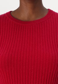 Roter gerippter Pullover mit rundem Halsausschnitt. Das strukturierte Strickmuster weist vertikale Streifen auf und hat ein weiches, warmes Aussehen.