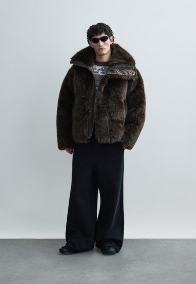 Giacca marrone in pelliccia sintetica con collo alto, zip frontale e grandi tasche. Abbinata a pantaloni neri a gamba larga e scarpe nere.