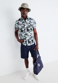 Camicia a maniche corte con bottoni, decorata con motivi di foglie nere su uno sfondo azzurro chiaro, abbinata a pantaloni corti di denim e una borsa tote blu navy.