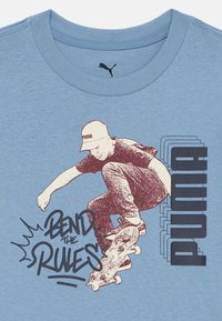Skateboarder iført caps som utfører et triks med teksten "Bend the Rules" og lagdelt "PUMA"-logo på en blå t-skjorte med rund hals.