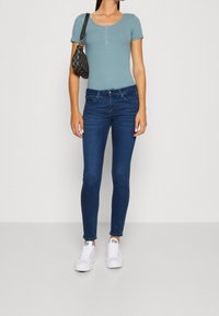 Top de canalé de manga corta en azul claro con detalle de botones, combinado con jeans ajustados en azul oscuro y zapatillas blancas, sostenido con un bolso negro.