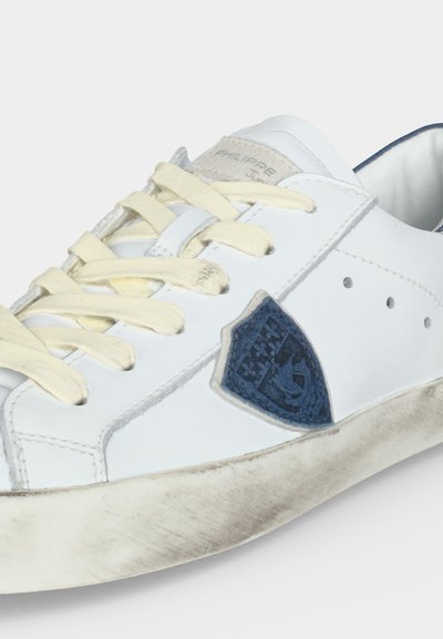 Baskets blanches avec des lacets jaunis, un logo blason bleu sur le côté et une semelle visiblement usée et sale sur un fond uni.