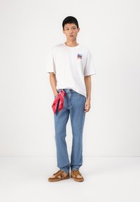 White cotton t-shirt with a small colorful graphic on the chest, paired with light blue denim jeans and a red bandana tied at the waist.