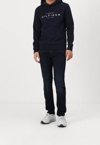 Uomo in felpa navy di Tommy Hilfiger, jeans scuri strappati e sneakers bianche in piedi contro uno sfondo chiaro.