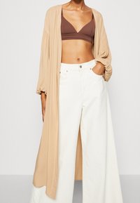 Beżowy, długi kardigan z luźnymi rękawami, połączony z brązową biżuterią i białymi spodniami z szerokimi nogawkami, ukazujący gładką teksturę materiału.