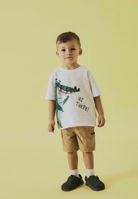 Una camiseta blanca de manga corta presenta un gráfico de un cocodrilo verde con el texto "¡HOLA!", combinada con pantalones cortos beige y zapatos negros. Fondo amarillo.