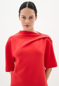 Röd oversized t-shirt av mjuk bomull. Har en unik draperad halsringning och korta ärmar. Enkelt design utan synliga mönster eller detaljer.