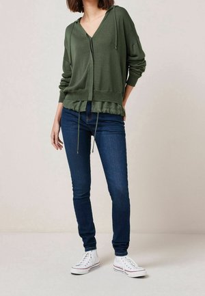 Felpa con cappuccio verde oliva, scollatura a V, dettagli con cordini e orlo arricciato, abbinata a jeans skinny blu scuro e sneakers bianche.