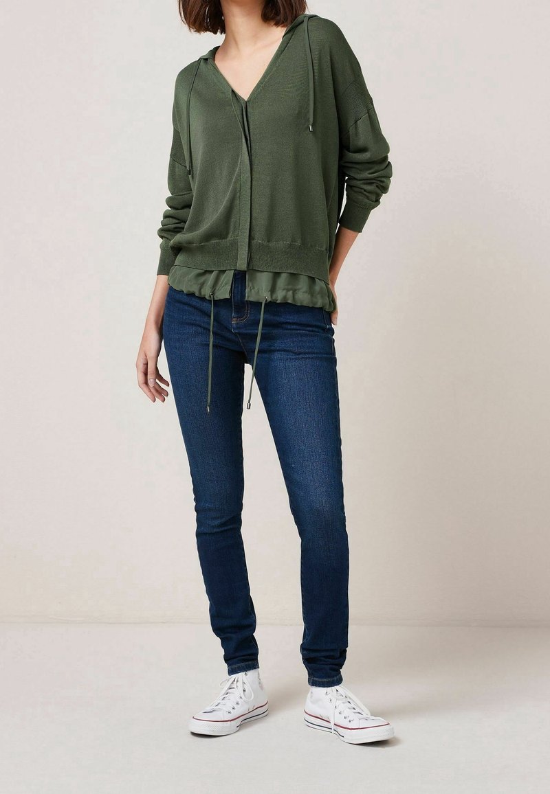 Olivgrünes Kapuzenshirt mit V-Ausschnitt, Kordelzugdetails und Rüschensaum, kombiniert mit dunkelblauen Skinny-Jeans und weißen Sneakers.