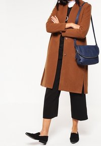 Kvinde iført en brun frakke, sorte cropped bukser, sorte loafers og en marineblå crossbody taske, stående med armene krydset.