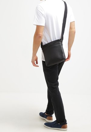 Černá kožená kabelka přes rameno s nastavitelným popruhem, obdélníkový tvar, texturovaný povrch a minimální kovové prvky, nošena osobou.