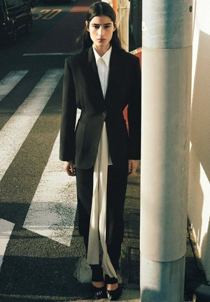 Frau mit langen dunklen Haaren, die einen schwarzen Anzug und ein weißes Hemd trägt, steht an einem städtischen Fußgängerüberweg bei Tageslicht in der Nähe eines Lichtmastes.