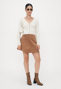 Witte geribde cardigan met knopen aan de voorkant, bruine suède A-lijnrok, bruine enkellaarzen en ronde groene zonnebril. Minimalistische handtas.
