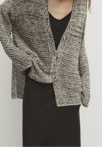 Neulecardigan beige- ja harmaanvärisenä, jossa on rakenteellinen kuosi, V-pääntie ja kolme nappia edessä. Päällä musta mekko.