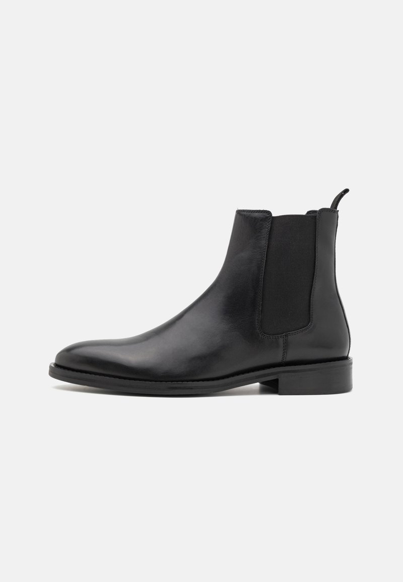 Botte Chelsea en cuir noir avec panneau latéral élastique, talon bas et languette de traction sur fond clair uni.
