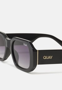Zwarte oversized zonnebril met gradient lenzen. Heeft opvallende vierkante monturen, goudaccenten op de armen en het "QUAY" logo aan de voorkant en zijkant.