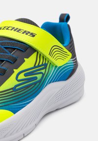 Baskets de sport avec un design noir, bleu et jaune fluo ; tissu texturé, sangle ajustable et semelle blanche avec un motif à pois.