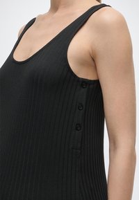 Czarny, bezrękawnik o prążkowanej fakturze z dekoltem w serek i trzema czarnymi guzikami wzdłuż bocznego szwu, noszony przez osobę o jasnej karnacji.