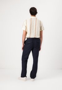 Korte mouwen shirt in crème met een bruin geometrisch patroon, gecombineerd met donkerblauwe broeken en witte sneakers, gezien vanuit de achterkant.