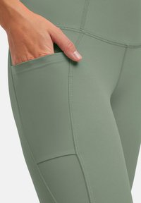 Leggings atletici verdi con una texture liscia, dotati di una tasca laterale e cuciture verticali per un design strutturato.