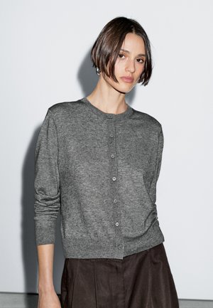 Woman with short dark hair wearing a buttoned grey cardigan and dark brown high-waisted pants against a plain white background.