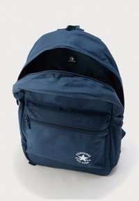 Σκούρο μπλε σακίδιο πλάτης Converse με εμπρόσθια τσέπη με φερμουάρ, λευκό λογότυπο και ανοιχτό κεντρικό διαμέρισμα σε λευκό φόντο.
