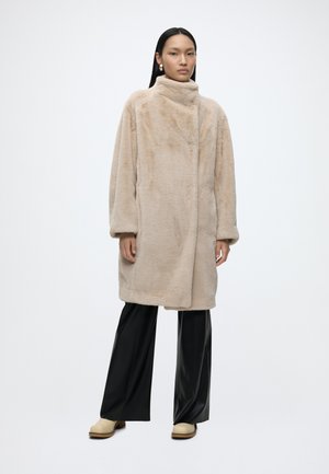 Frau mit langen schwarzen Haaren, die einen beigen Kunstpelzmantel, schwarze Hosen mit weitem Bein, beige Schuhe und goldene Ohrringe trägt, steht vor weißem Hintergrund.