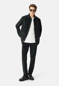 Giacca in corduroy nera con chiusura a bottoni, dotata di una tasca sul petto, abbinata a pantaloni a sigaretta coordinati e scarpe nere con lacci.