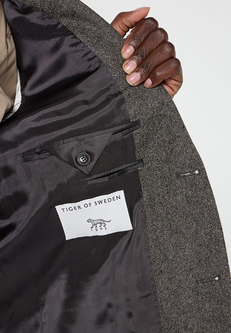 Grauer Blazer-Innenbereich mit schwarzem Futter, ausgestattet mit einer Tasche, Knopfdetails und einem "TIGER OF SWEDEN"-Etikett mit einem Tiger-Graphic.