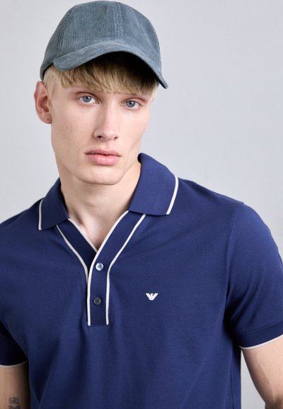 Tricou polo bleu cu dungi albe, două butoane și un mic logo pe piept. Căciulă din catifea gri. Modelul are părul blond deschis și ochii albaștri.