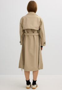 Gabardina color beige con un cuello desmontable, cintura ceñida con cinturón y mangas largas. Textura suave y diseño de longitud hasta la rodilla. El calzado incluye calcetas negras y zapatillas deportivas.
