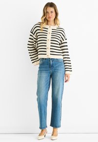 Striped knit cardigan in beige and black with large buttons, paired with light blue straight-leg jeans and cream block-heel shoes.