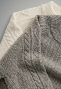 Twee truien: één crème met een gestructureerd kabelbreipatroon, de andere grijs met een vergelijkbaar kabelpatroon. Beide hebben geribbelde kragen en een zachte, pluizige textuur.
