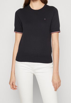 Vrouw die een zwart kortärmelig truitje met een klein logo op de borst en een witte broek draagt, staand tegen een effen lichte achtergrond.