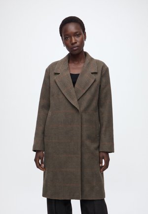 Femme portant un long manteau marron à carreaux avec de larges revers par-dessus un haut noir, debout devant un fond blanc uni.