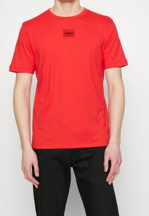 Homme portant un T-shirt rouge uni à manches courtes avec un patch logo "HUGO" noir sur la poitrine, associé à un pantalon noir sur fond blanc uni.
