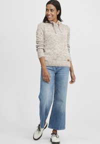 Beige strikkegenser med hette og en treknapp, kombinert med lyseblå vide jeans og hvite sko med svarte snører.