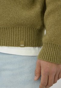 Pull tricoté vert olive avec poignets et ourlet côtelés. Présente une étiquette en tissu avec le logo "BZE". Porté sur une chemise blanche et un jean bleu.