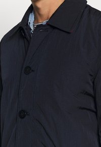 Marineblaue gesteppte Jacke mit klassischem Kragen, schwarzen Knöpfen und glatter Textur; weist einen dezenten Glanz und ein minimalistisches Design auf.