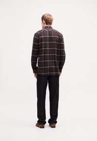Mann, der ein braun-schwarz karriertes Langarmhemd, schwarze Hosen und braune Schuhe trägt, steht mit dem Gesicht von der Kamera abgewandt vor einem schlichten weißen Hintergrund.