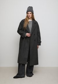 Cappotto di lana grigio con design doppiopetto, grandi rever e tasche laterali, abbinato a un maglione grigio e pantaloni jeans a gamba larga scuri.
