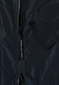 Gros plan sur une veste bleu marine foncé montrant une fermeture éclair noire avec un tire-zip marqué "CECIL OUTERWEAR" et une manche avec détail de sangle en Velcro.