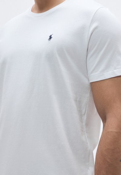 Fehér pamutpóló, kerek nyakkivágással és rövid ujjal, a bal mellkasrészen kis tengerészkék logóval.