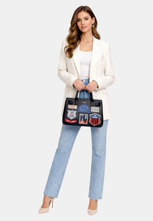 Žena oblečená v bílém saku, bílém topu, světle modrých džínách a béžových podpatcích, držící černou kabelku s barevnými nášivkami s tématem Paříže.