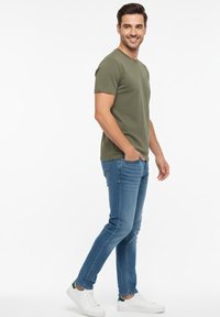 Camiseta de manga corta color verde oliva combinada con jeans azules y zapatillas blancas con detalles en verde, posando sobre un fondo blanco liso.